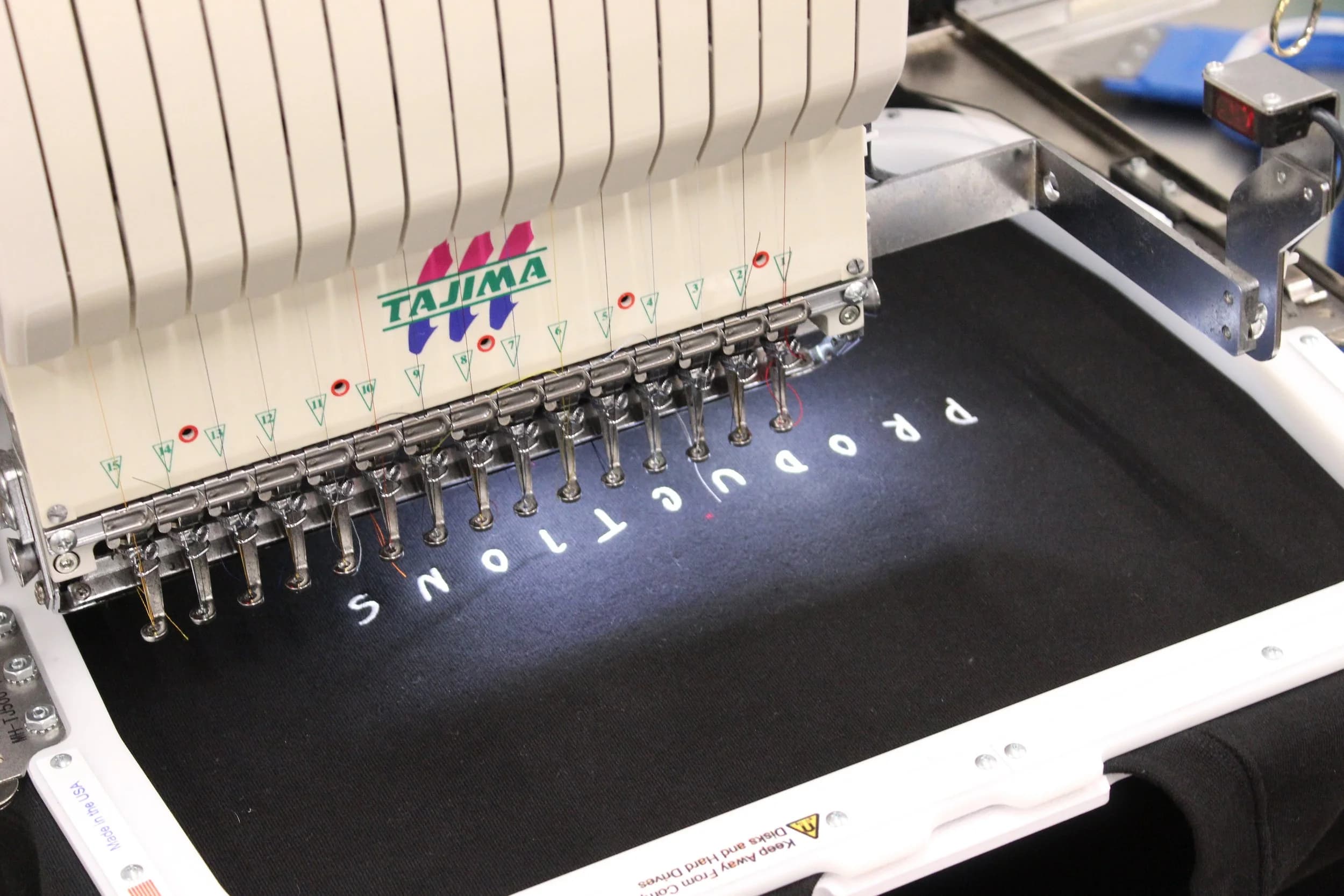 A multi-needle embroidery machine stitching the Eminence wordmark into a black garment