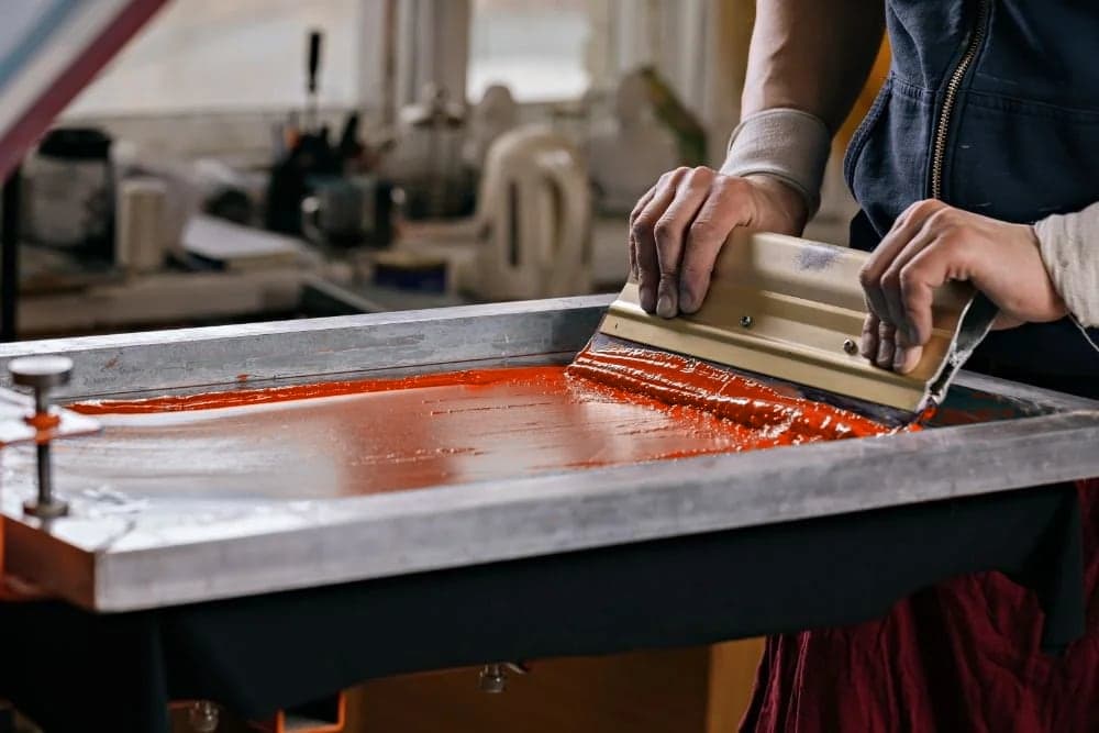 A squeegee pulling orange ink across a screen on a printing press
